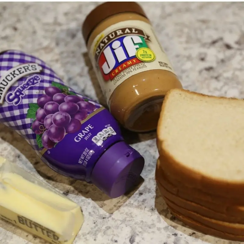 The Ultimate Grilled Peanut Butter and Jelly Sandwich Recipe 13 Ingredients for a grilled peanut butter and jelly sandwich laid out on a kitchen countertop, featuring four slices of white bread, a jar of creamy Jif peanut butter, a jar of Smucker's grape jelly, and a full stick of butter.