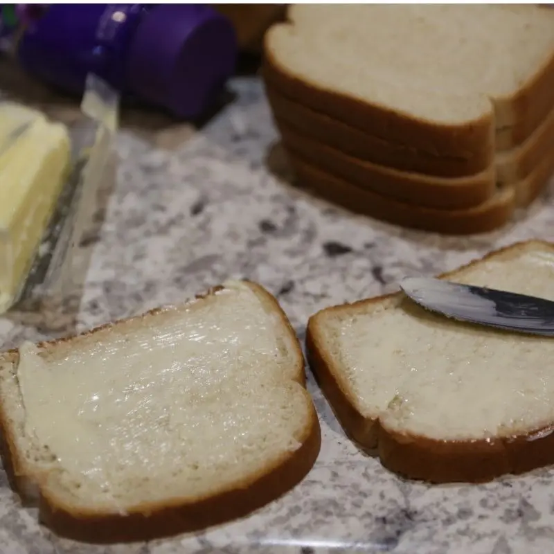 The Ultimate Grilled Peanut Butter and Jelly Sandwich Recipe 14 Two slices of white bread on a blue plate, each topped with a dollop of room-temperature butter. A silver butter knife sits nearby, ready to spread the butter for grilling.