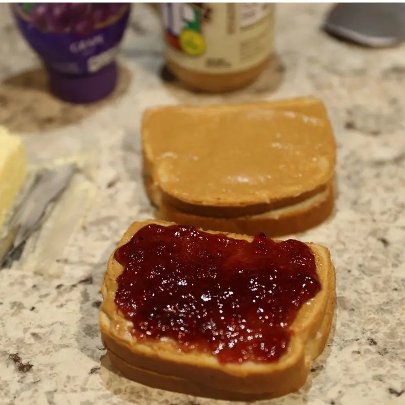 The Ultimate Grilled Peanut Butter and Jelly Sandwich Recipe 16 Two slices of bread for a grilled peanut butter and jelly sandwich. Both slices are coated with peanut butter, and one slice has a generous layer of dark purple Smucker’s grape jelly spread over it, ready to be closed and grilled.
