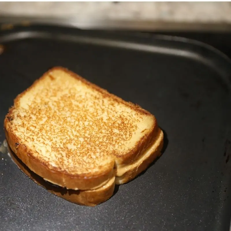 The Ultimate Grilled Peanut Butter and Jelly Sandwich Recipe 17 A perfectly toasted, golden-brown grilled peanut butter and jelly sandwich on a frying pan. The bread has a buttery crisp texture with warm peanut butter beginning to melt at the edges.