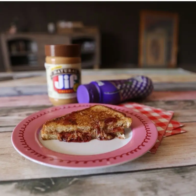 The Ultimate Grilled Peanut Butter and Jelly Sandwich Recipe 19 A close-up of half a grilled peanut butter and jelly sandwich on a pink and white plate, with melted Jif peanut butter and Smucker's grape jelly oozing from the center. In the blurred background, a jar of Jif Natural peanut butter and Smucker's grape jelly sit on the table.