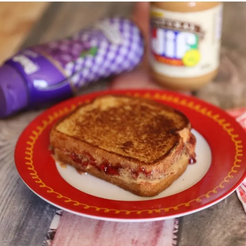 The Ultimate Grilled Peanut Butter and Jelly Sandwich Recipe 10 A perfectly toasted, golden-brown grilled peanut butter and jelly sandwich sliced in half on a red plate, showing the warm, melted Jif Natural peanut butter and gooey Smucker's grape jelly oozing from the sides.