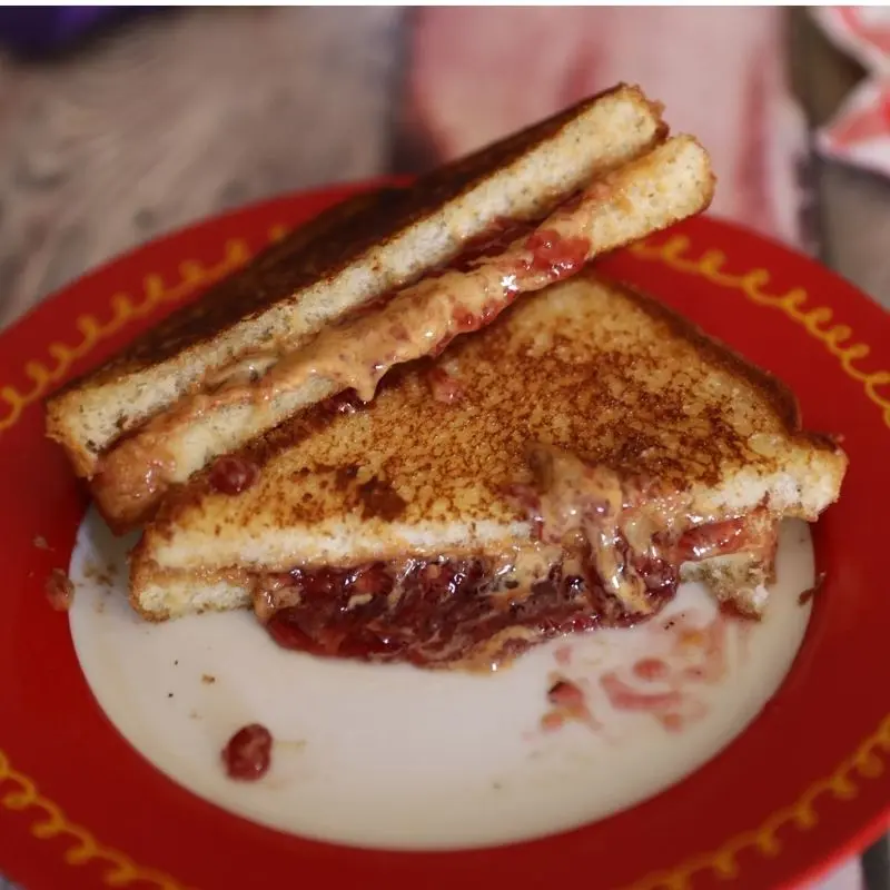 The Ultimate Grilled Peanut Butter and Jelly Sandwich Recipe 11 A messy, delicious grilled peanut butter and jelly sandwich sliced in half on a red plate. The golden-brown toasted bread is filled with melted Jif peanut butter and Smucker's grape jelly that is oozing out from the center and dripping down the sides.